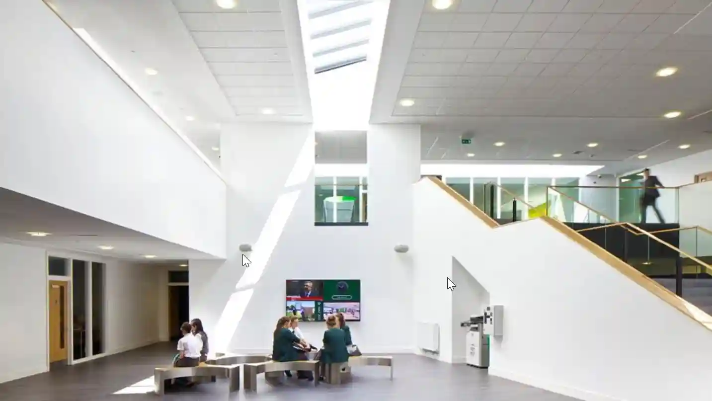 Bright and spacious school atrium with white walls, a high ceiling with skylights, a wide staircase, and students seated at tables.