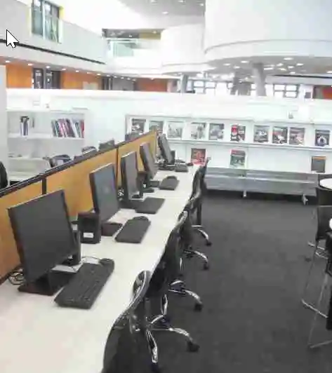 School library with rows of computers on desks, bookshelves with colorful book covers, and a modern interior design featuring a two-story layout with balconies.